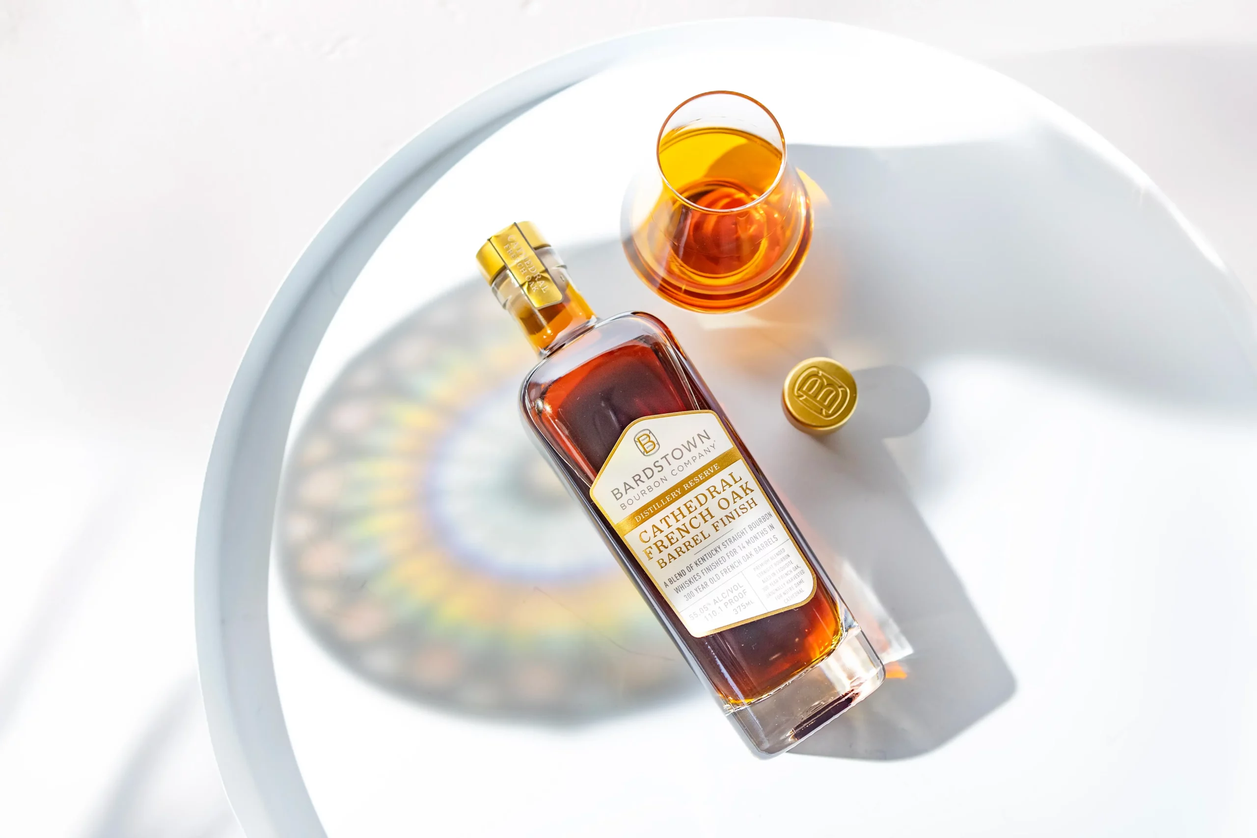 Bardstown Bourbon Cathedral French Oak Barrel Finish bottle beside a glass, stained-glass reflection on the table
