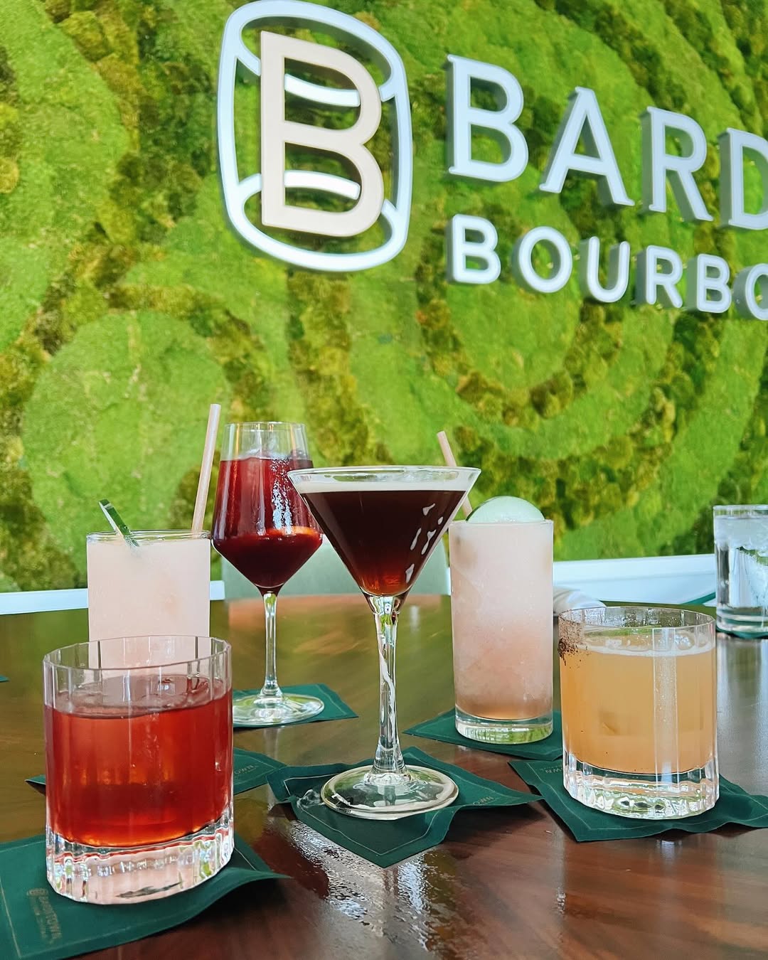 Colorful bourbon cocktails displayed on a wooden table in front of Bardstown Bourbon’s vibrant green moss wall logo.