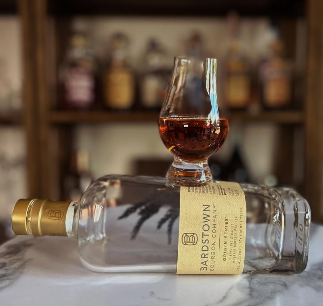 Bardstown Bourbon Origin Series bottle on marble with a Glencairn glass of bourbon, bar shelf blurred in the background.