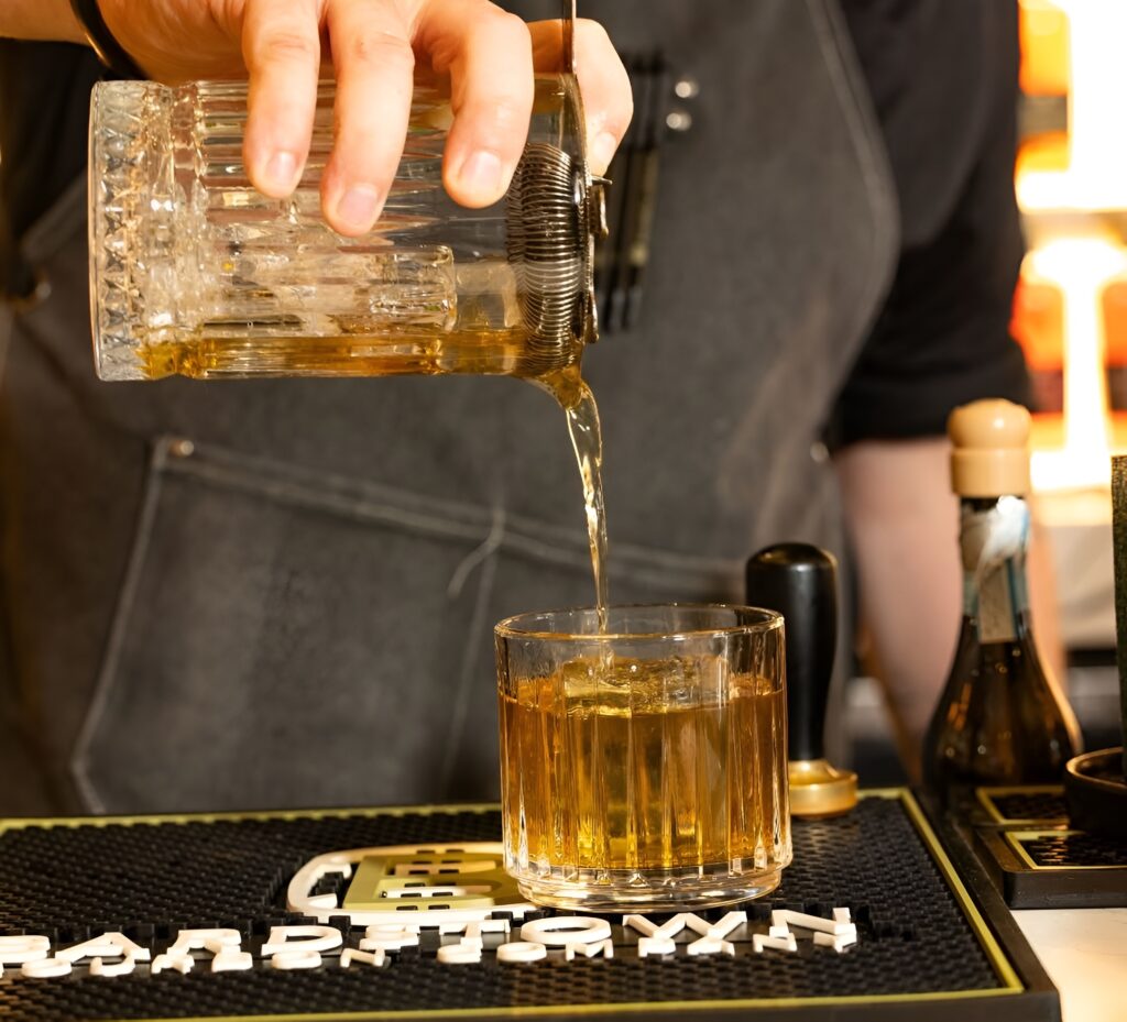 Bartender pouring a golden Bardstown Bourbon cocktail with precision, set against a branded bar mat.