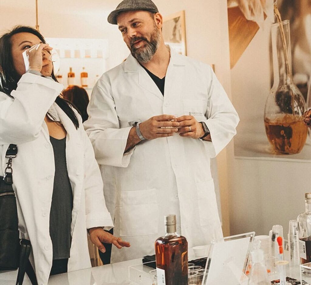 Two people in lab coats sampling whiskey at a Bardstown Bourbon blending experience inside a modern tasting room.