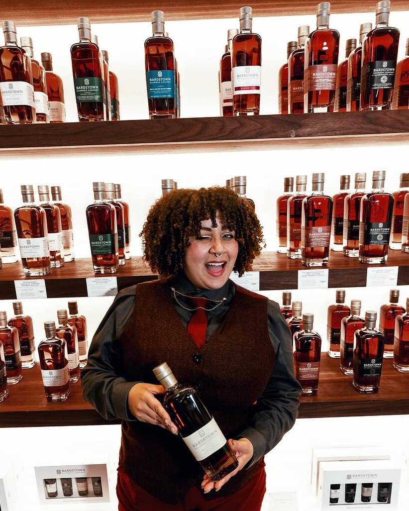 Smiling staff member holding a Bardstown Bourbon bottle in front of a display wall filled with Bardstown Bourbon.