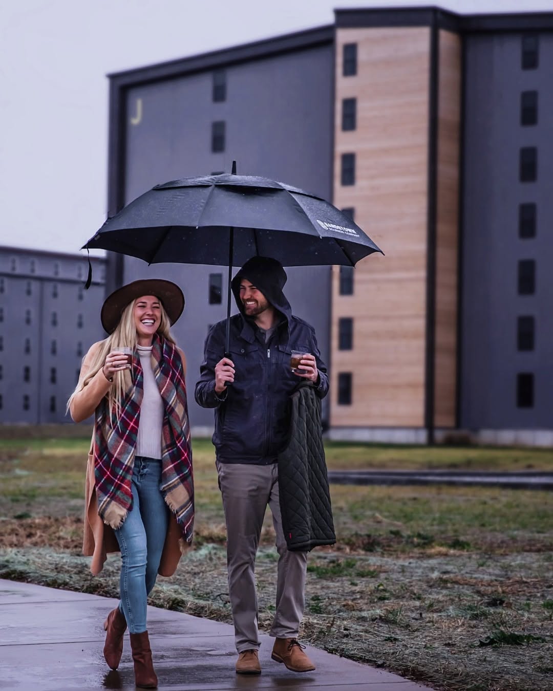 Couple walking under an umbrella near Bardstown Bourbon Company distillery, enjoying drinks on a rainy day.